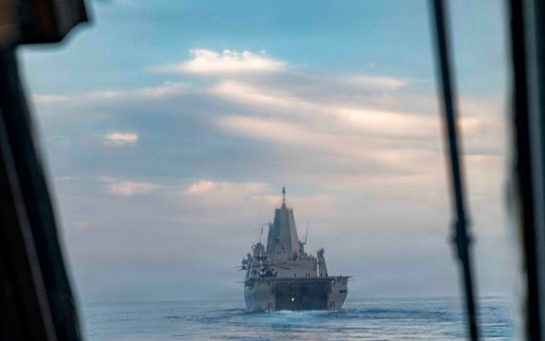 The image is taken from the cockpit of a military aircraft or helicopter, looking through the windscreen at a large naval vessel underway at sea. A person in the foreground, likely a pilot or crew member, is partially visible on the left side. The naval vessel appears to be a US Navy amphibious transport dock (LPD-class) ship, viewed from the bow/front aspect.