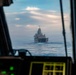 The image is taken from the cockpit of a military aircraft or helicopter, looking through the windscreen at a large naval vessel underway at sea. A person in the foreground, likely a pilot or crew member, is partially visible on the left side. The naval vessel appears to be a US Navy amphibious transport dock (LPD-class) ship, viewed from the bow/front aspect.