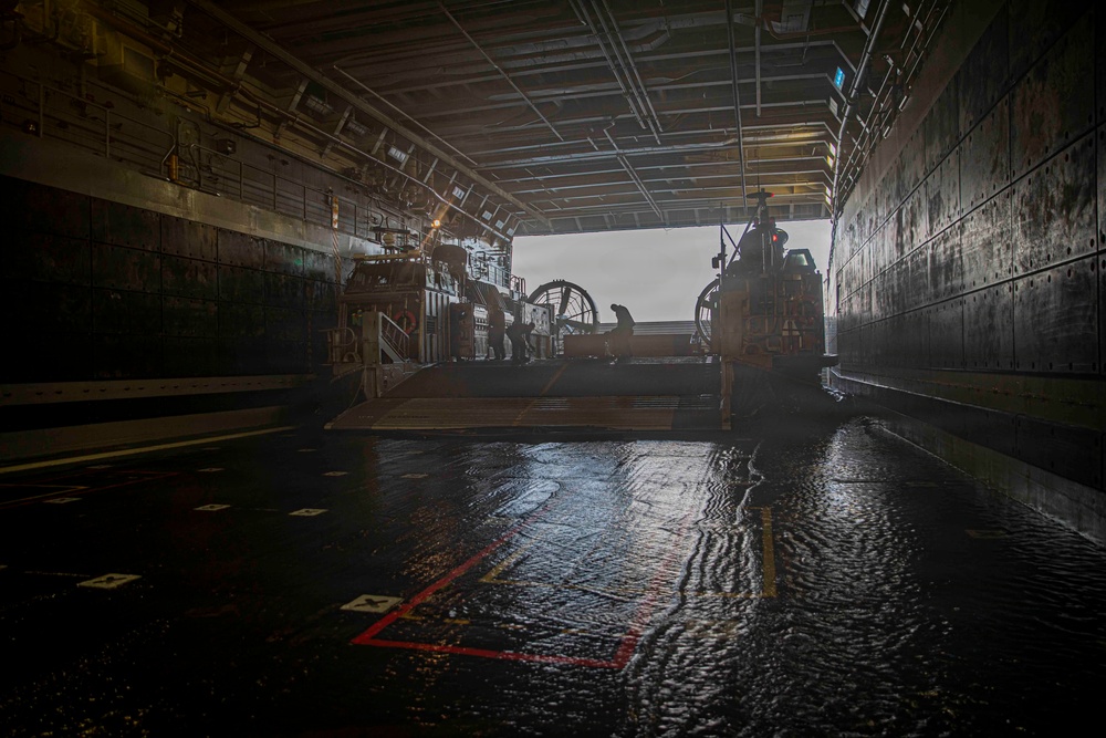 The image shows the interior of a naval vessel's well deck, with a Landing Craft Air Cushion (LCAC) or similar amphibious craft positioned inside. The wet, reflective deck floor and open stern gate reveal overcast conditions outside. The space is characterized by industrial infrastructure including overhead piping, lighting, and marked deck zones.