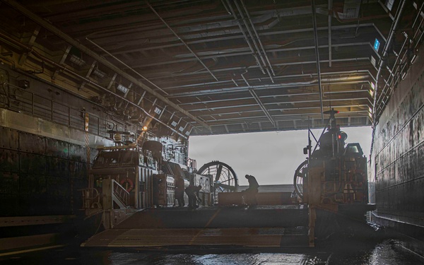 The image shows the interior of a naval vessel's well deck, with a Landing Craft Air Cushion (LCAC) or similar amphibious craft positioned inside. The wet, reflective deck floor and open stern gate reveal overcast conditions outside. The space is characterized by industrial infrastructure including overhead piping, lighting, and marked deck zones.