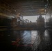 The image shows the interior of a naval vessel's well deck, with a Landing Craft Air Cushion (LCAC) or similar amphibious craft positioned inside. The wet, reflective deck floor and open stern gate reveal overcast conditions outside. The space is characterized by industrial infrastructure including overhead piping, lighting, and marked deck zones.