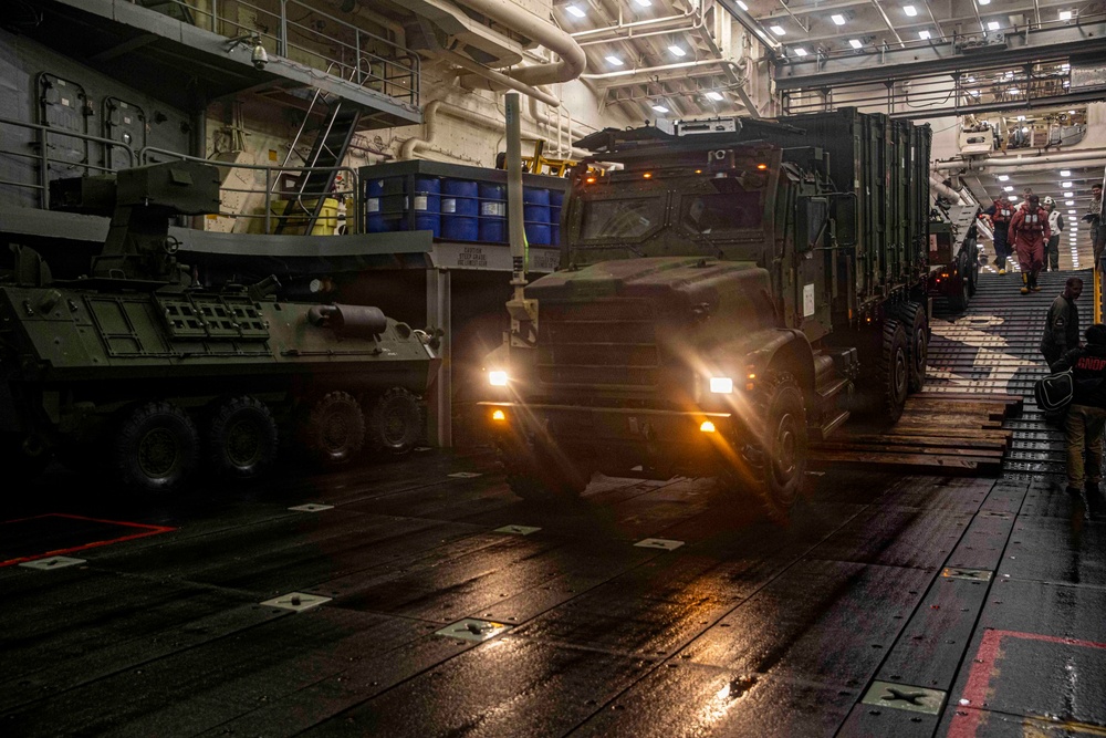 Military vehicles are being loaded or unloaded inside the well deck of a naval amphibious vessel. A large military tactical truck (appearing to be an FMTV or similar medium tactical vehicle) is driving through the dimly lit interior, alongside what appears to be a wheeled armored vehicle on the left. Personnel in red and dark uniforms are visible guiding the operation.