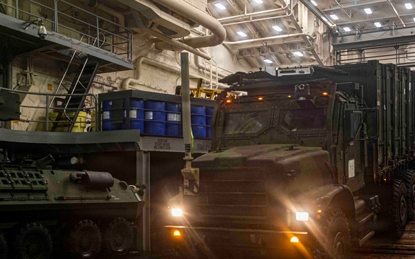 Military vehicles are being loaded or unloaded inside the well deck of a naval amphibious vessel. A large military tactical truck (appearing to be an FMTV or similar medium tactical vehicle) is driving through the dimly lit interior, alongside what appears to be a wheeled armored vehicle on the left. Personnel in red and dark uniforms are visible guiding the operation.