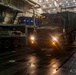 Military vehicles are being loaded or unloaded inside the well deck of a naval amphibious vessel. A large military tactical truck (appearing to be an FMTV or similar medium tactical vehicle) is driving through the dimly lit interior, alongside what appears to be a wheeled armored vehicle on the left. Personnel in red and dark uniforms are visible guiding the operation.