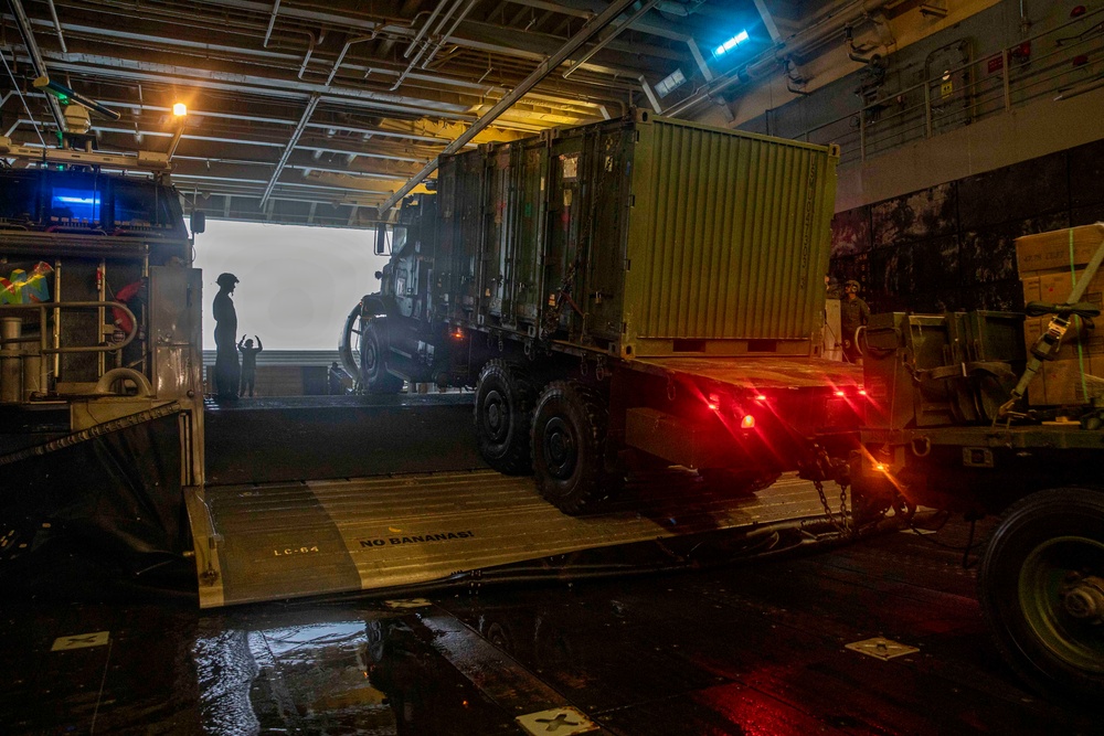 A military vehicle loaded with a green cargo container is being driven down a ramp from the well deck of a naval vessel, likely a landing ship or amphibious assault ship. The scene is dimly lit with red tail lights illuminating the ramp area and a bright opening visible at the stern. At least one silhouetted personnel is visible directing or observing the offloading operation.