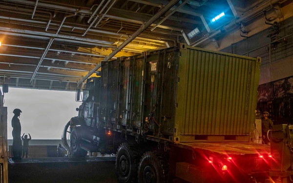 A military vehicle loaded with a green cargo container is being driven down a ramp from the well deck of a naval vessel, likely a landing ship or amphibious assault ship. The scene is dimly lit with red tail lights illuminating the ramp area and a bright opening visible at the stern. At least one silhouetted personnel is visible directing or observing the offloading operation.