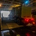 A military vehicle loaded with a green cargo container is being driven down a ramp from the well deck of a naval vessel, likely a landing ship or amphibious assault ship. The scene is dimly lit with red tail lights illuminating the ramp area and a bright opening visible at the stern. At least one silhouetted personnel is visible directing or observing the offloading operation.