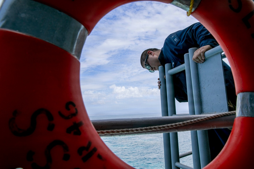 A person in a dark blue uniform leans over a ship's railing, viewed through the opening of a red life preserver ring mounted on the vessel's side. The composition frames the subject dramatically through the life ring, with open ocean and partly cloudy sky visible in the background. The image appears to depict a maritime crew member conducting an inspection or observation over the side of the vessel.