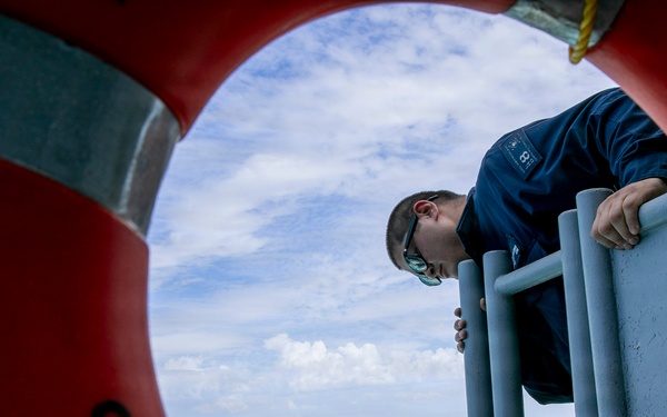 A person in a dark blue uniform leans over a ship's railing, viewed through the opening of a red life preserver ring mounted on the vessel's side. The composition frames the subject dramatically through the life ring, with open ocean and partly cloudy sky visible in the background. The image appears to depict a maritime crew member conducting an inspection or observation over the side of the vessel.