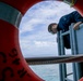 A person in a dark blue uniform leans over a ship's railing, viewed through the opening of a red life preserver ring mounted on the vessel's side. The composition frames the subject dramatically through the life ring, with open ocean and partly cloudy sky visible in the background. The image appears to depict a maritime crew member conducting an inspection or observation over the side of the vessel.