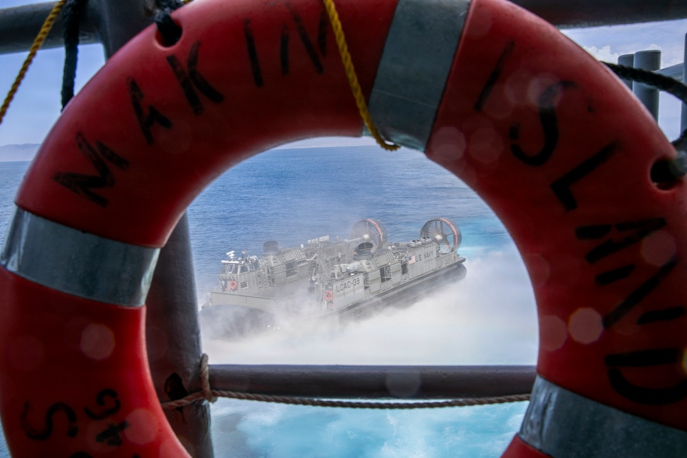 A U.S. Navy Landing Craft Air Cushion (LCAC) hovercraft, designated LCAC-33, is photographed through the opening of a red life preserver ring mounted on a ship's railing. The life ring bears partial text reading 'MARIN' and 'ISLAND' with markings 'S.S. 24'. The hovercraft is moving at speed across open water, generating a large spray of mist and foam.