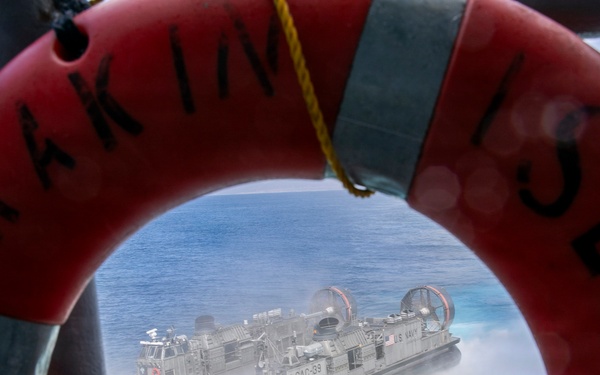 A U.S. Navy Landing Craft Air Cushion (LCAC) hovercraft, designated LCAC-33, is photographed through the opening of a red life preserver ring mounted on a ship's railing. The life ring bears partial text reading 'MARIN' and 'ISLAND' with markings 'S.S. 24'. The hovercraft is moving at speed across open water, generating a large spray of mist and foam.