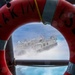 A U.S. Navy Landing Craft Air Cushion (LCAC) hovercraft, designated LCAC-33, is photographed through the opening of a red life preserver ring mounted on a ship's railing. The life ring bears partial text reading 'MARIN' and 'ISLAND' with markings 'S.S. 24'. The hovercraft is moving at speed across open water, generating a large spray of mist and foam.
