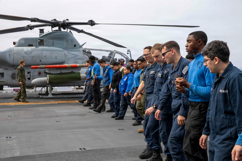 A group of U.S. Navy personnel and individuals in civilian/mixed attire are lined up on the flight deck of a naval vessel, appearing to observe or receive a briefing near a parked military helicopter. A Marine Corps AH-1Z Viper attack helicopter is prominently visible on the deck with orange safety covers on its weapons pylons. The overcast sky and open ocean are visible in the background, confirming an at-sea setting.