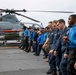 A group of U.S. Navy personnel and individuals in civilian/mixed attire are lined up on the flight deck of a naval vessel, appearing to observe or receive a briefing near a parked military helicopter. A Marine Corps AH-1Z Viper attack helicopter is prominently visible on the deck with orange safety covers on its weapons pylons. The overcast sky and open ocean are visible in the background, confirming an at-sea setting.