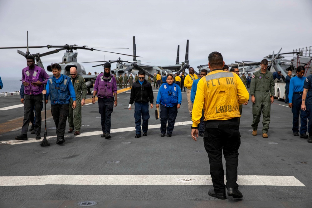 Personnel are conducting a foreign object debris (FOD) walkdown on the flight deck of a U.S. Navy amphibious assault ship, identifiable by 'FLT DK LHD-2' stenciled on the back of a yellow jersey worn by a flight deck officer. Crew members in various colored jerseys and flight suits walk in a line across the deck, scanning for debris. Military helicopters and tiltrotor aircraft (MV-22 Ospreys and AH-1Z Vipers) are visible in the background.