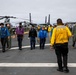 Personnel are conducting a foreign object debris (FOD) walkdown on the flight deck of a U.S. Navy amphibious assault ship, identifiable by 'FLT DK LHD-2' stenciled on the back of a yellow jersey worn by a flight deck officer. Crew members in various colored jerseys and flight suits walk in a line across the deck, scanning for debris. Military helicopters and tiltrotor aircraft (MV-22 Ospreys and AH-1Z Vipers) are visible in the background.