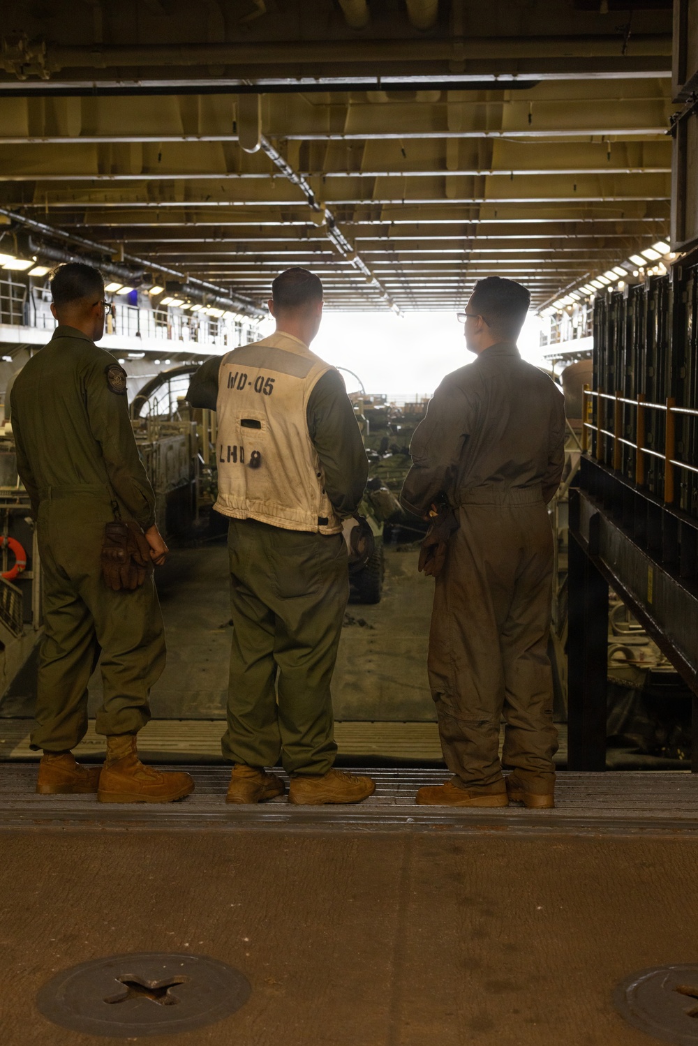 Three military personnel stand inside what appears to be the well deck of a naval amphibious assault ship, identified as LHD-8 (USS Makin Island) based on the vest markings. The central figure wears a white working vest marked 'WD-05 LHD 8', indicating a well deck crew role. The personnel are facing toward the open stern gate, with military vehicles and equipment visible in the background.