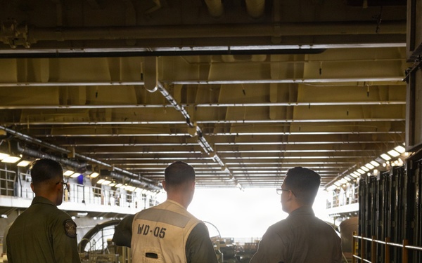 Three military personnel stand inside what appears to be the well deck of a naval amphibious assault ship, identified as LHD-8 (USS Makin Island) based on the vest markings. The central figure wears a white working vest marked 'WD-05 LHD 8', indicating a well deck crew role. The personnel are facing toward the open stern gate, with military vehicles and equipment visible in the background.