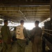 Three military personnel stand inside what appears to be the well deck of a naval amphibious assault ship, identified as LHD-8 (USS Makin Island) based on the vest markings. The central figure wears a white working vest marked 'WD-05 LHD 8', indicating a well deck crew role. The personnel are facing toward the open stern gate, with military vehicles and equipment visible in the background.