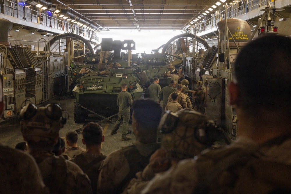 Military personnel are gathered inside what appears to be the well deck of a naval amphibious assault ship, surrounding multiple armored vehicles secured with chains. Soldiers wearing combat gear, helmets, and hearing protection are positioned around the vehicles, suggesting a loading or staging operation. The interior structure shows overhead lighting, catwalks, and safety signage consistent with a Landing Craft Air Cushion (LCAC) or similar amphibious vessel well deck.