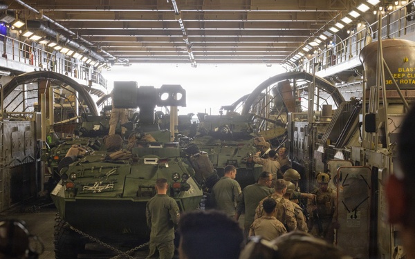 Military personnel are gathered inside what appears to be the well deck of a naval amphibious assault ship, surrounding multiple armored vehicles secured with chains. Soldiers wearing combat gear, helmets, and hearing protection are positioned around the vehicles, suggesting a loading or staging operation. The interior structure shows overhead lighting, catwalks, and safety signage consistent with a Landing Craft Air Cushion (LCAC) or similar amphibious vessel well deck.