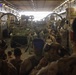 Military personnel are gathered inside what appears to be the well deck of a naval amphibious assault ship, surrounding multiple armored vehicles secured with chains. Soldiers wearing combat gear, helmets, and hearing protection are positioned around the vehicles, suggesting a loading or staging operation. The interior structure shows overhead lighting, catwalks, and safety signage consistent with a Landing Craft Air Cushion (LCAC) or similar amphibious vessel well deck.