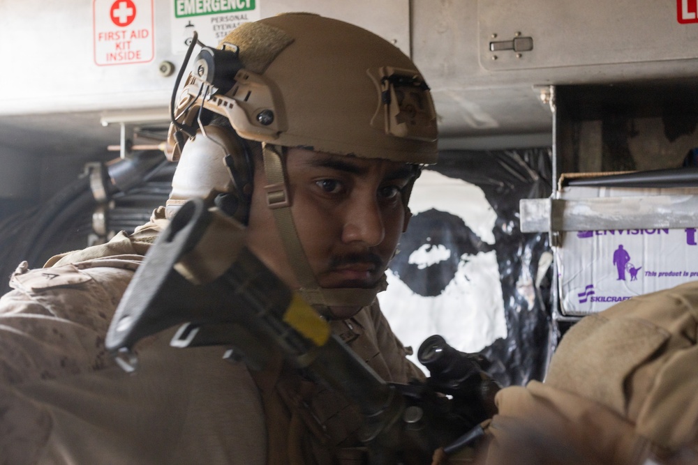 A military service member wearing tan/coyote brown combat gear and a ballistic helmet with communications equipment is positioned inside what appears to be a military vehicle or facility. The individual is holding a weapon and appears to be in a tactical posture, looking alert. Safety signage including a First Aid Kit and Emergency Eyewash station are visible in the background.