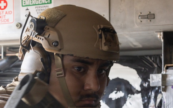 A military service member wearing tan/coyote brown combat gear and a ballistic helmet with communications equipment is positioned inside what appears to be a military vehicle or facility. The individual is holding a weapon and appears to be in a tactical posture, looking alert. Safety signage including a First Aid Kit and Emergency Eyewash station are visible in the background.