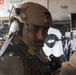 A military service member wearing tan/coyote brown combat gear and a ballistic helmet with communications equipment is positioned inside what appears to be a military vehicle or facility. The individual is holding a weapon and appears to be in a tactical posture, looking alert. Safety signage including a First Aid Kit and Emergency Eyewash station are visible in the background.