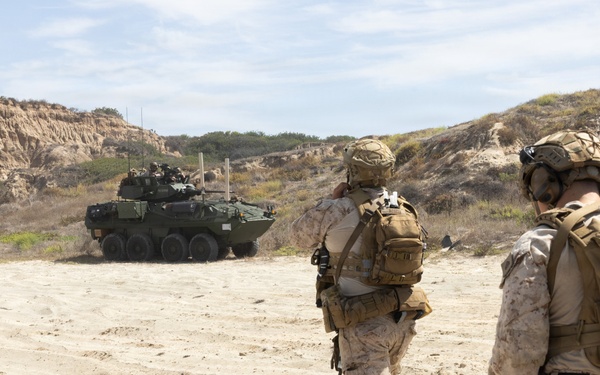 Three military personnel in desert camouflage uniforms and tactical gear are walking across a sandy terrain toward a Light Armored Vehicle (LAV), likely an LAV-25 or Stryker-type wheeled armored vehicle. The setting appears to be a coastal training area with sandy bluffs and scrub vegetation visible in the background. The scene is consistent with a military training exercise.