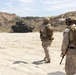Three military personnel in desert camouflage uniforms and tactical gear are walking across a sandy terrain toward a Light Armored Vehicle (LAV), likely an LAV-25 or Stryker-type wheeled armored vehicle. The setting appears to be a coastal training area with sandy bluffs and scrub vegetation visible in the background. The scene is consistent with a military training exercise.