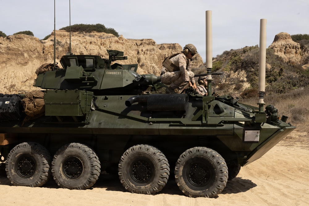 A U.S. Marine Corps Light Armored Vehicle (LAV-25 or variant) named 'CONQUEST' is positioned on sandy terrain with eroded sandstone bluffs in the background. Two personnel in desert camouflage uniforms and helmets are visible on top of the vehicle, appearing to conduct maintenance or operational checks. The vehicle is an 8-wheeled armored fighting vehicle equipped with a turret, multiple antennas, and external gear.