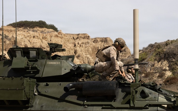 A U.S. Marine Corps Light Armored Vehicle (LAV-25 or variant) named 'CONQUEST' is positioned on sandy terrain with eroded sandstone bluffs in the background. Two personnel in desert camouflage uniforms and helmets are visible on top of the vehicle, appearing to conduct maintenance or operational checks. The vehicle is an 8-wheeled armored fighting vehicle equipped with a turret, multiple antennas, and external gear.
