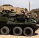 A U.S. Marine Corps Light Armored Vehicle (LAV-25 or variant) named 'CONQUEST' is positioned on sandy terrain with eroded sandstone bluffs in the background. Two personnel in desert camouflage uniforms and helmets are visible on top of the vehicle, appearing to conduct maintenance or operational checks. The vehicle is an 8-wheeled armored fighting vehicle equipped with a turret, multiple antennas, and external gear.