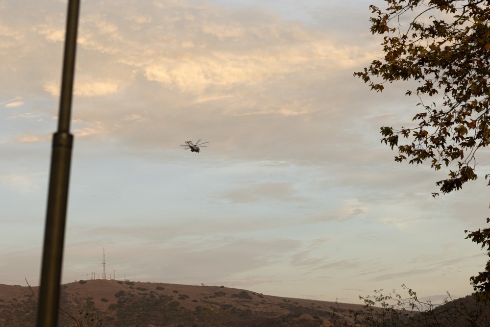A helicopter is captured in flight over a hilly, arid landscape during what appears to be late afternoon or dusk, given the warm golden light on the clouds. The scene includes communication towers or antennas visible on a distant ridgeline. The foreground features a blurred vertical pole on the left and tree branches on the right.