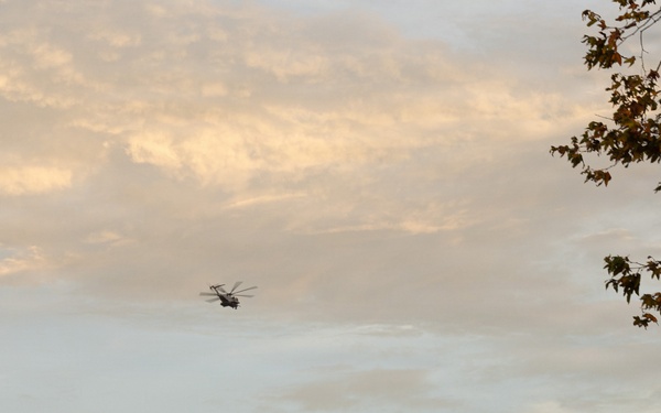 A helicopter is captured in flight over a hilly, arid landscape during what appears to be late afternoon or dusk, given the warm golden light on the clouds. The scene includes communication towers or antennas visible on a distant ridgeline. The foreground features a blurred vertical pole on the left and tree branches on the right.