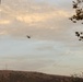 A helicopter is captured in flight over a hilly, arid landscape during what appears to be late afternoon or dusk, given the warm golden light on the clouds. The scene includes communication towers or antennas visible on a distant ridgeline. The foreground features a blurred vertical pole on the left and tree branches on the right.