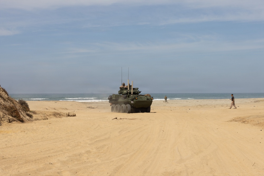 A military wheeled armored vehicle, consistent with an LAV-25 or Stryker-type 8-wheeled armored personnel carrier, is traversing a sandy beach near the ocean shoreline. At least one crew member is visible in the open hatch, and several personnel are present in the surrounding area. The scene appears to be a military training exercise or amphibious landing drill.