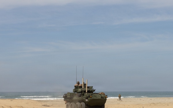 A military wheeled armored vehicle, consistent with an LAV-25 or Stryker-type 8-wheeled armored personnel carrier, is traversing a sandy beach near the ocean shoreline. At least one crew member is visible in the open hatch, and several personnel are present in the surrounding area. The scene appears to be a military training exercise or amphibious landing drill.