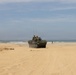A military wheeled armored vehicle, consistent with an LAV-25 or Stryker-type 8-wheeled armored personnel carrier, is traversing a sandy beach near the ocean shoreline. At least one crew member is visible in the open hatch, and several personnel are present in the surrounding area. The scene appears to be a military training exercise or amphibious landing drill.