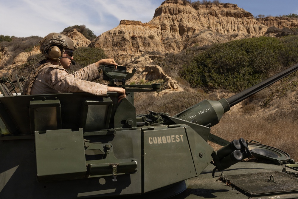 A U.S. Marine in desert MARPAT camouflage and combat helmet is positioned in the open hatch of a military armored vehicle named 'CONQUEST', operating in a rugged, arid terrain with eroded sandstone cliffs in the background. The vehicle appears to be an LAV-25 or similar light armored vehicle equipped with a cannon. The setting suggests a training exercise, likely at a base in Southern California based on the distinctive terrain.