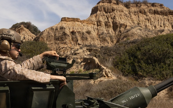 A U.S. Marine in desert MARPAT camouflage and combat helmet is positioned in the open hatch of a military armored vehicle named 'CONQUEST', operating in a rugged, arid terrain with eroded sandstone cliffs in the background. The vehicle appears to be an LAV-25 or similar light armored vehicle equipped with a cannon. The setting suggests a training exercise, likely at a base in Southern California based on the distinctive terrain.