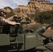 A U.S. Marine in desert MARPAT camouflage and combat helmet is positioned in the open hatch of a military armored vehicle named 'CONQUEST', operating in a rugged, arid terrain with eroded sandstone cliffs in the background. The vehicle appears to be an LAV-25 or similar light armored vehicle equipped with a cannon. The setting suggests a training exercise, likely at a base in Southern California based on the distinctive terrain.