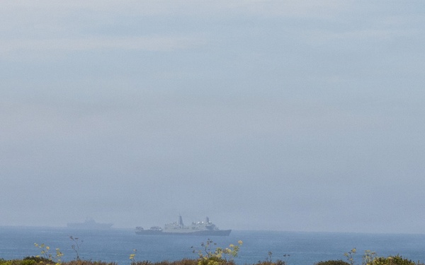 A coastal scene photographed from a low dune or scrubland area, showing a hazy ocean horizon with what appears to be one or more large naval or military vessels visible in the distance. The foreground is dominated by dry scrub vegetation typical of a Mediterranean or coastal chaparral environment. Atmospheric haze or sea fog significantly reduces visibility of the ships.