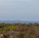A coastal scene photographed from a low dune or scrubland area, showing a hazy ocean horizon with what appears to be one or more large naval or military vessels visible in the distance. The foreground is dominated by dry scrub vegetation typical of a Mediterranean or coastal chaparral environment. Atmospheric haze or sea fog significantly reduces visibility of the ships.