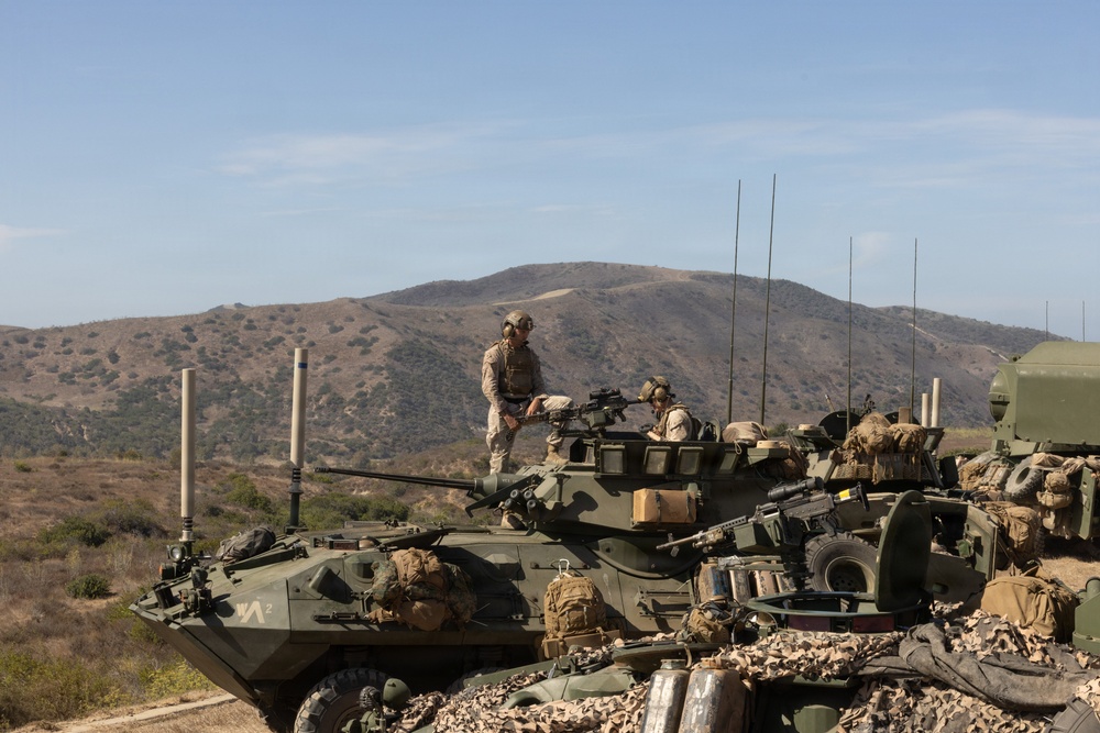 Military personnel in desert camouflage uniforms are positioned atop what appear to be LAV-25 (Light Armored Vehicle) armored vehicles during what appears to be a training exercise. The scene is set in an arid, hilly terrain consistent with a military training installation in the southwestern United States. Multiple vehicles are visible with various equipment, antennas, and gear loaded on them.