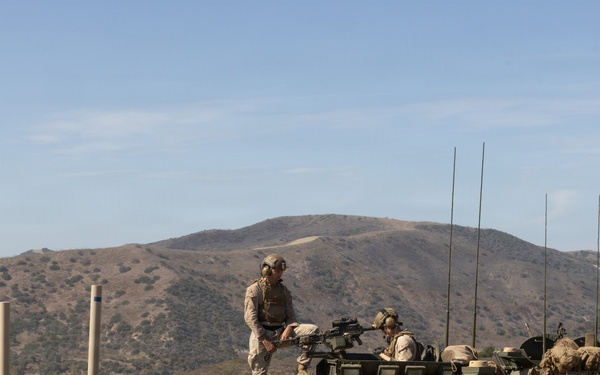 Military personnel in desert camouflage uniforms are positioned atop what appear to be LAV-25 (Light Armored Vehicle) armored vehicles during what appears to be a training exercise. The scene is set in an arid, hilly terrain consistent with a military training installation in the southwestern United States. Multiple vehicles are visible with various equipment, antennas, and gear loaded on them.