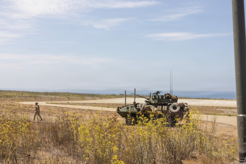 A military exercise or operation is taking place in a coastal terrain environment. An armored wheeled vehicle, consistent with a Light Armored Vehicle (LAV) or similar 8-wheeled armored personnel carrier, is positioned on or near a paved road with personnel visible around it. A single soldier in camouflage uniform is walking nearby, with the ocean visible in the background.