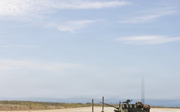 A military exercise or operation is taking place in a coastal terrain environment. An armored wheeled vehicle, consistent with a Light Armored Vehicle (LAV) or similar 8-wheeled armored personnel carrier, is positioned on or near a paved road with personnel visible around it. A single soldier in camouflage uniform is walking nearby, with the ocean visible in the background.